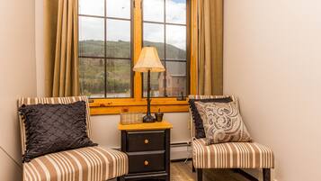 Two striped chairs with decorative pillows, a small wooden nightstand with a lamp, and a window with a view of a forested landscape.