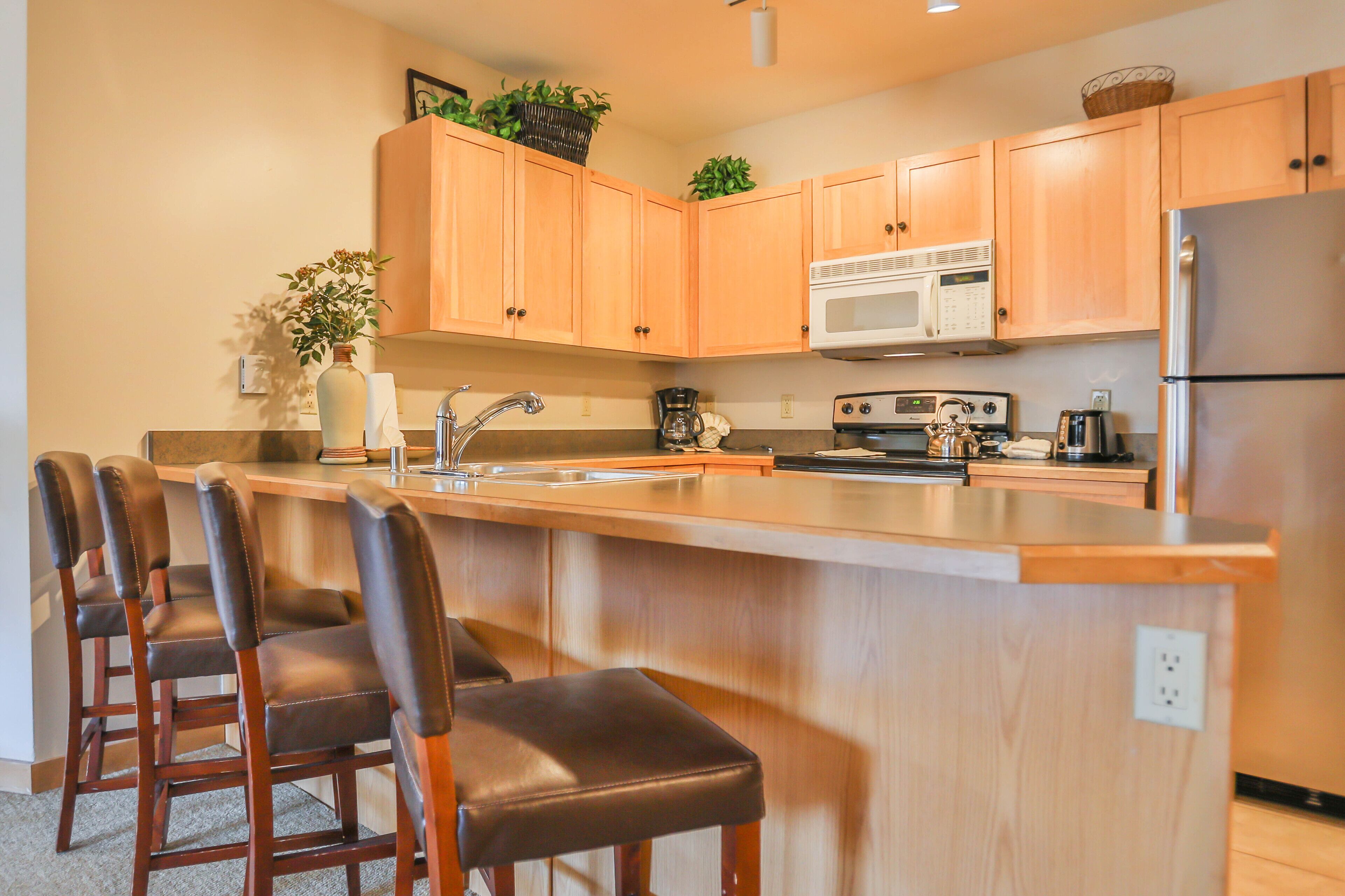 A kitchen with wooden cabinets, a stainless steel refrigerator, a microwave, and a coffee maker. There are four bar stools with leather seats.