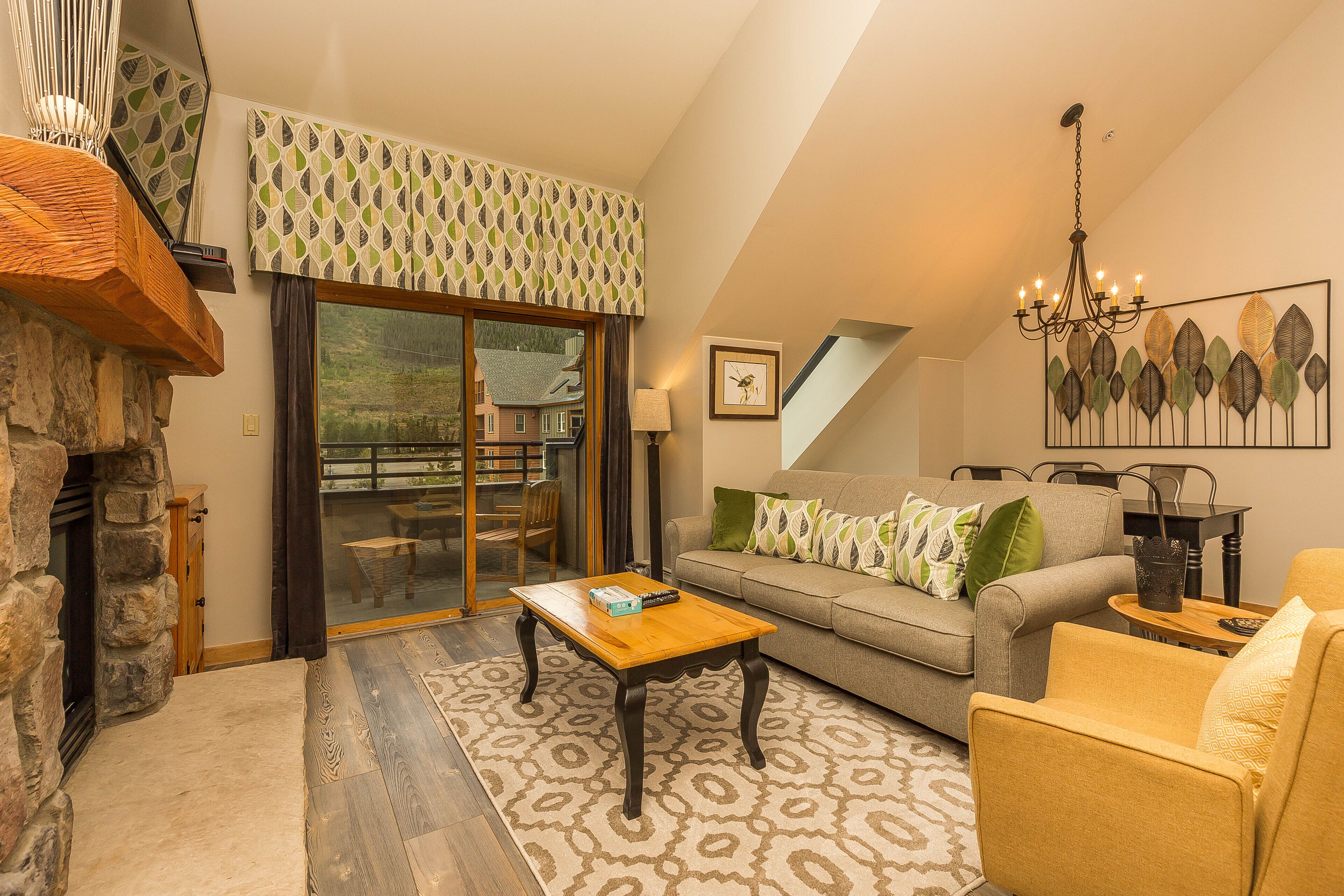 A cozy living room with a stone fireplace, a wooden coffee table, a sofa with patterned cushions, and a chandelier.
