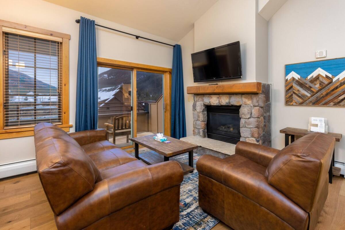A cozy living room with leather sofas, a stone fireplace, a flat-screen TV, and a large window with blue curtains.