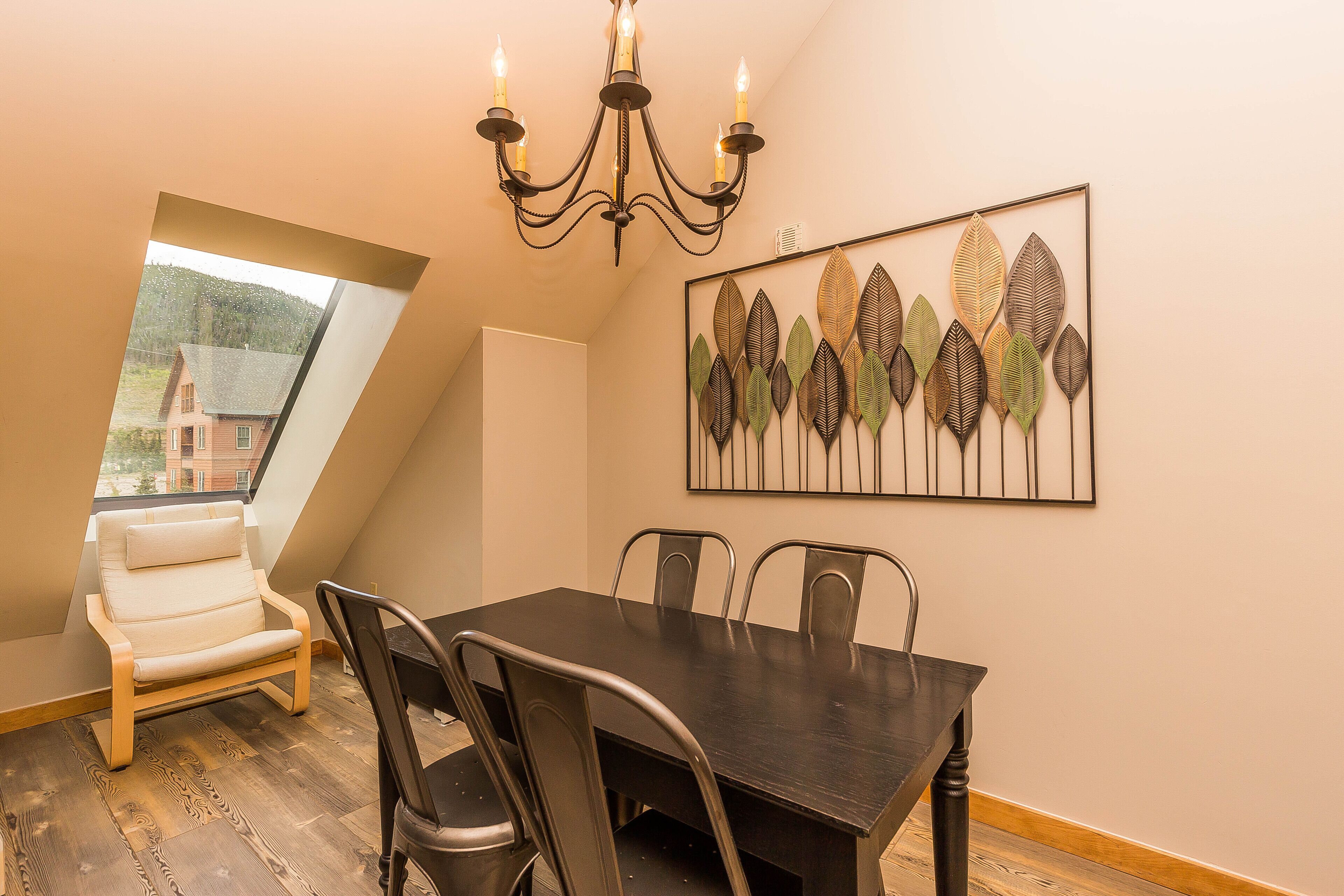 A dining area with a dark wooden table, four chairs, a white cushioned chair, a chandelier, and a leaf-patterned wall art.