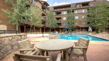 A pool area with wooden deck chairs and a table, surrounded by multi-story buildings with balconies and trees.