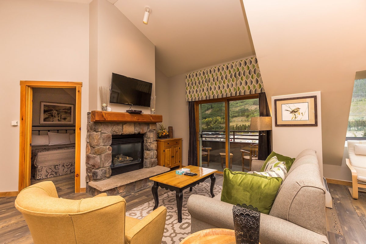 A living room with a stone fireplace, a television mounted on the wall, a sofa, a coffee table, and a view of the outdoors through a large window.