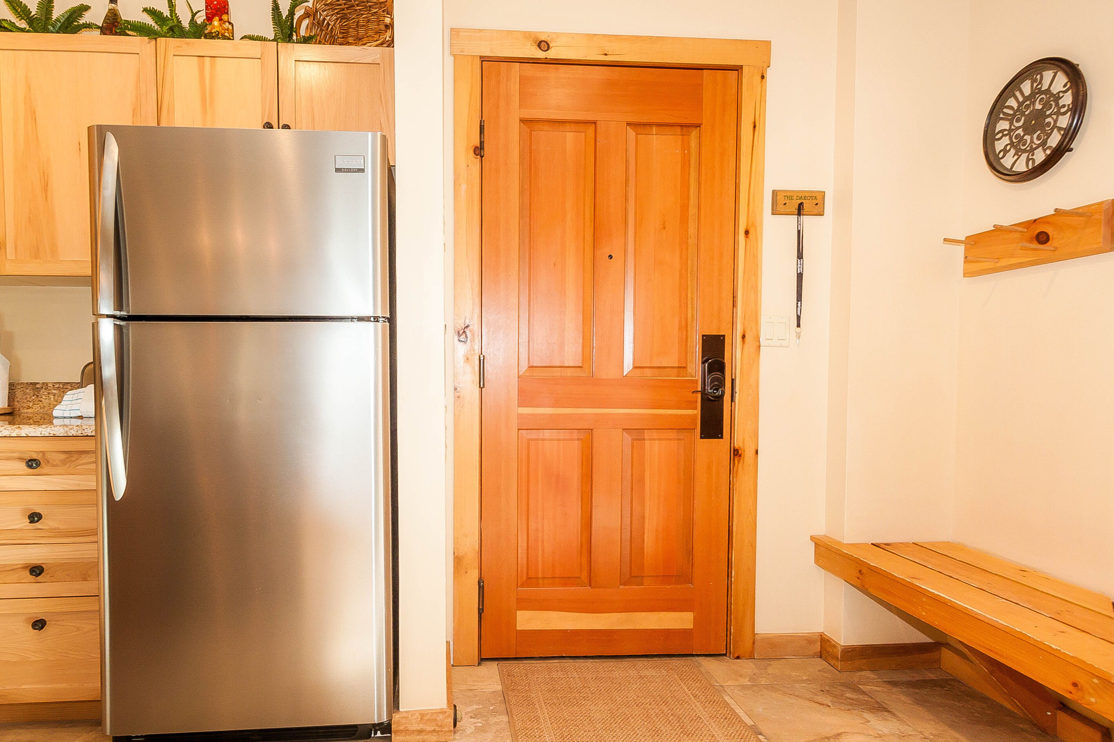 A kitchen with a stainless steel refrigerator, wooden cabinets, a wooden door, a wall clock, and a bench.