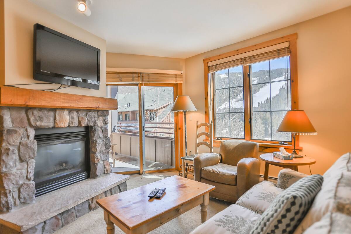 A cozy living room with a stone fireplace, a flat-screen TV mounted above it, a comfortable sofa, a coffee table, and a view of a snowy landscape through large windows.