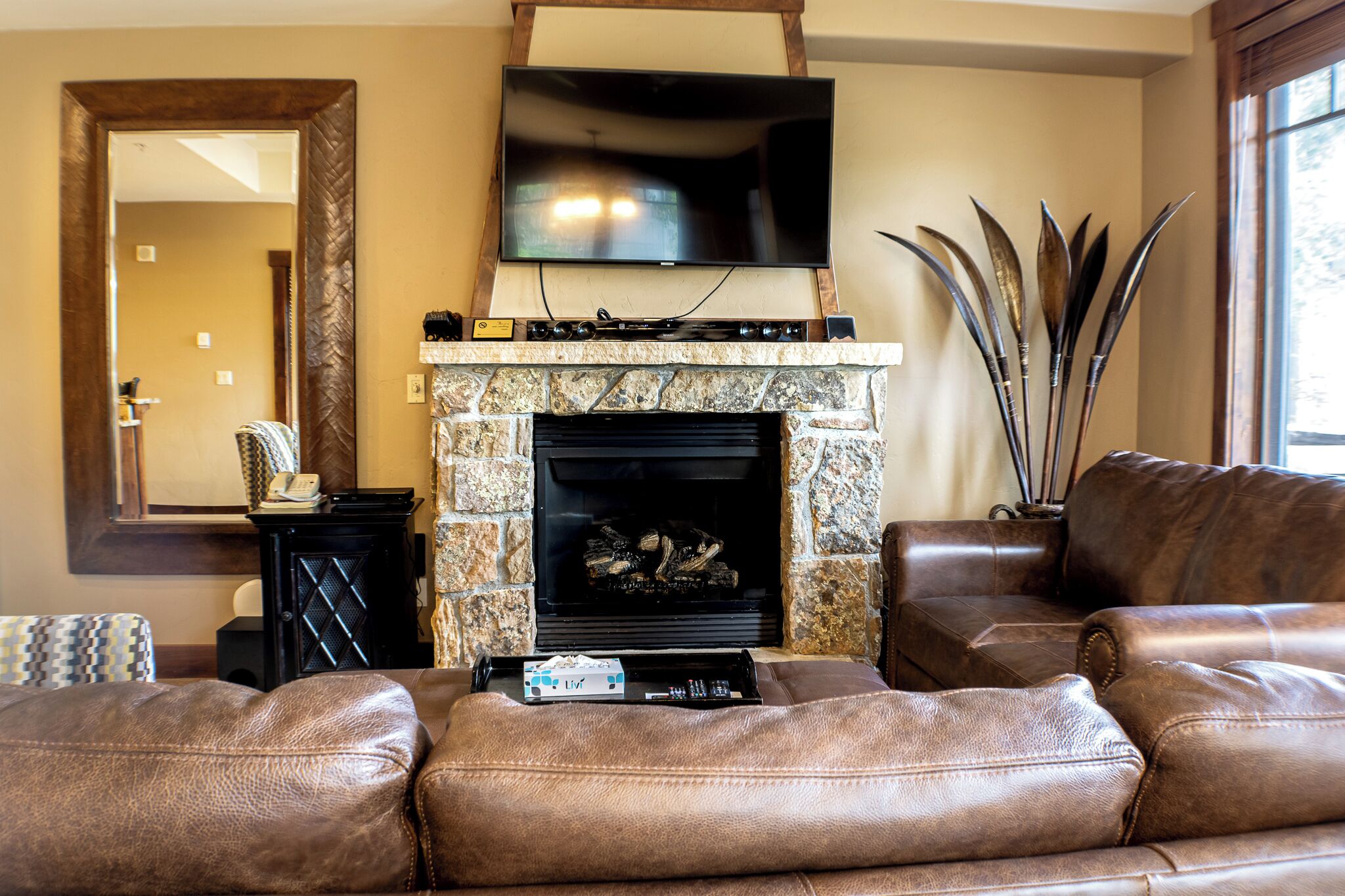 A living room with a stone fireplace, leather sofas, a flat-screen TV, and a large mirror.