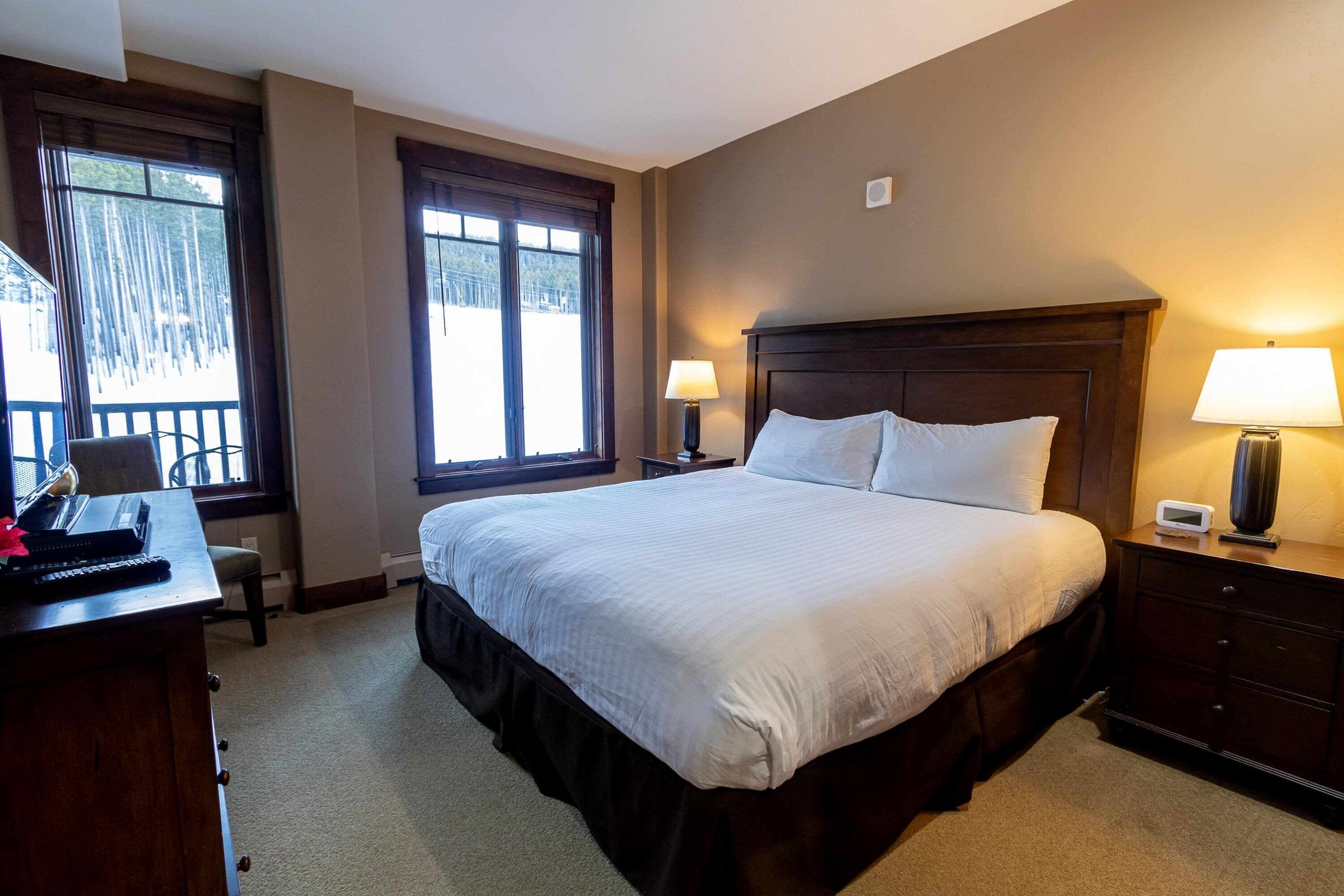 A hotel room with a large bed, a desk with a computer, a chair, a nightstand with a lamp, and a window with a view of trees.