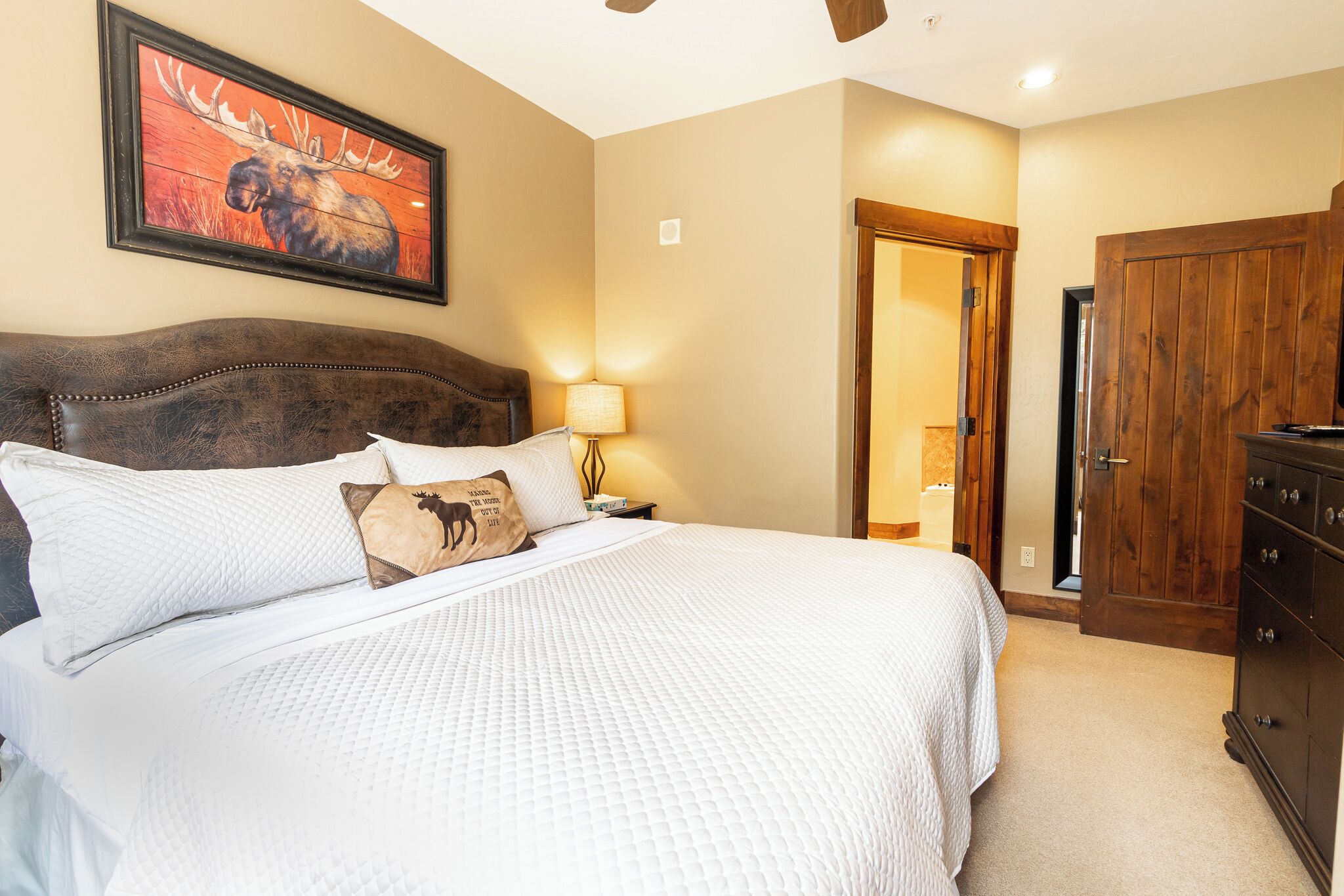 Un dormitorio con una cama grande, cabecera de madera, mesita de noche con lámpara, armario de madera y una puerta que da a otra habitación.