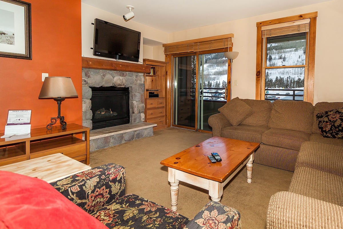 A living room with a stone fireplace, a television mounted above it, and a large window with a view of a snowy landscape.