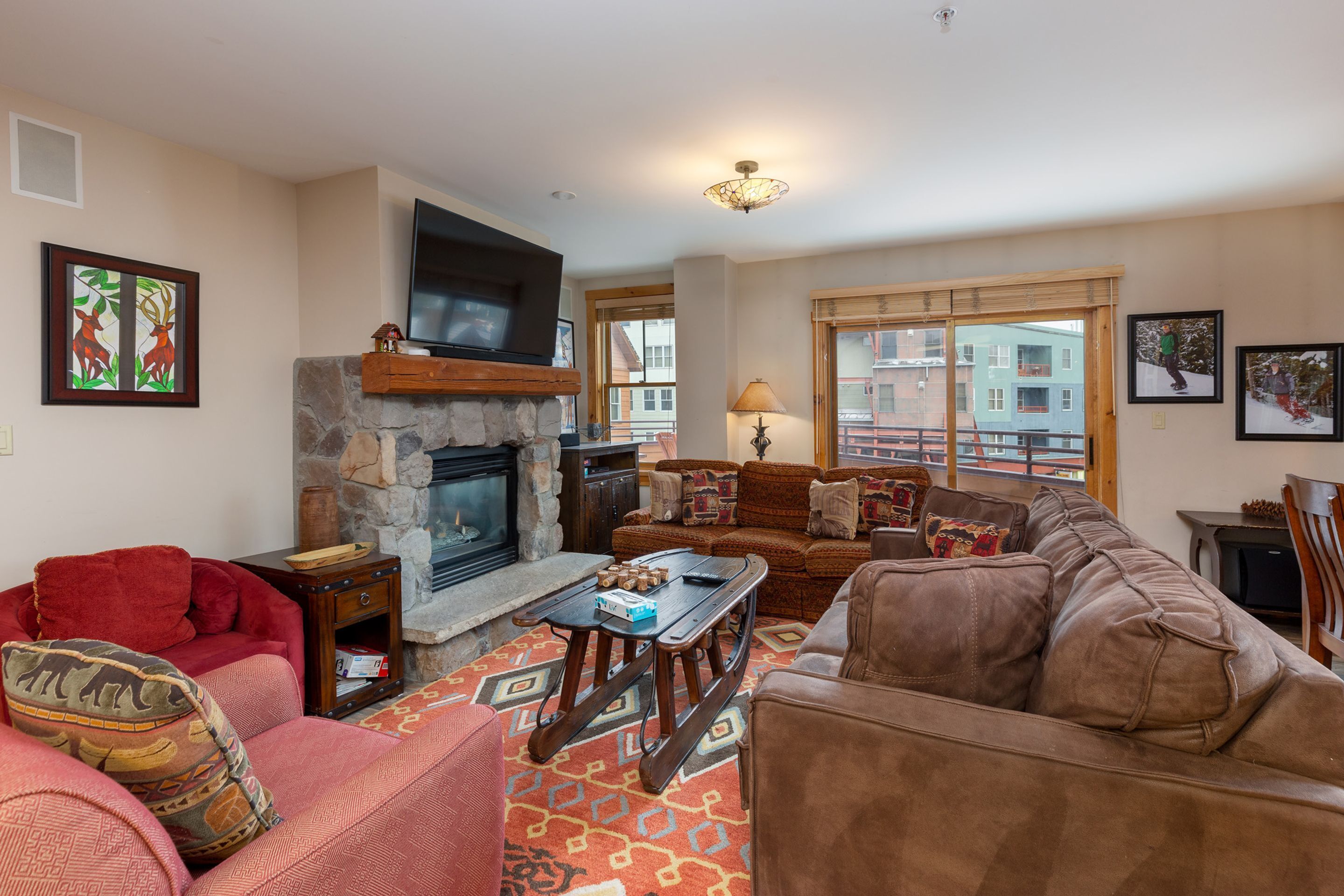 A cozy living room with a stone fireplace, a television mounted above it, and a large window offering a view of buildings outside.