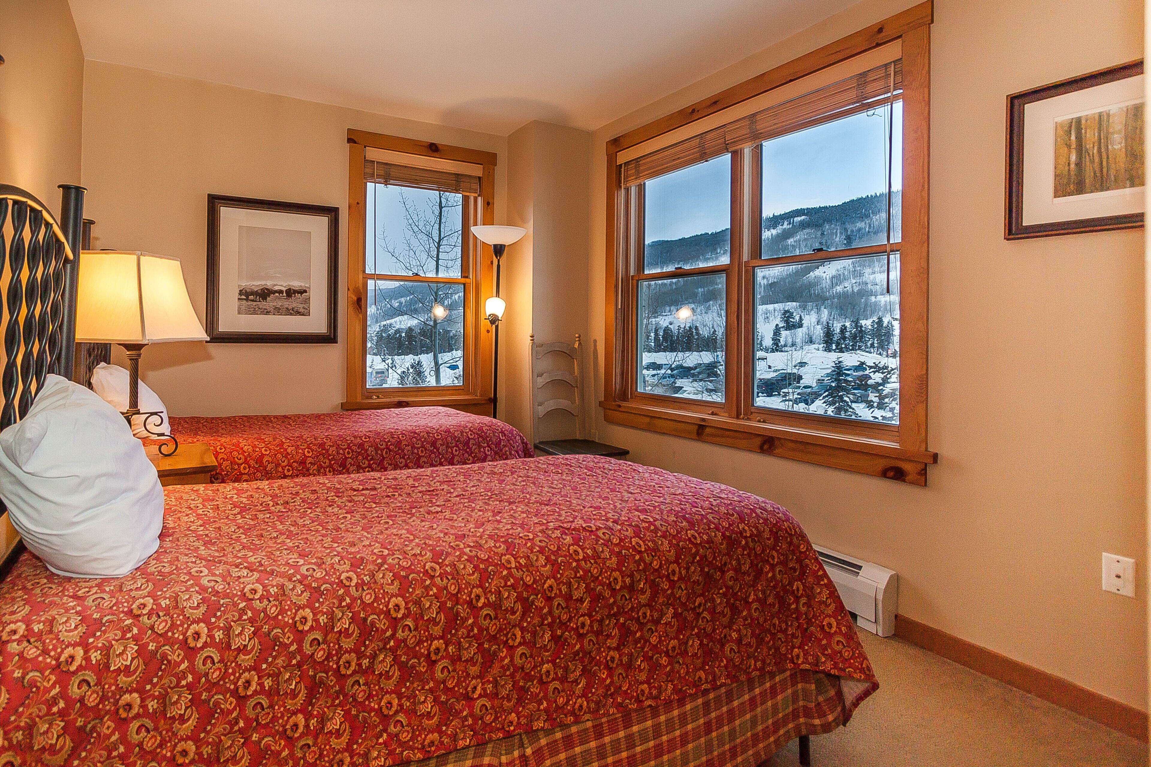 Un dormitorio con dos camas, una ventana que ofrece vistas nevadas, una lámpara y un cuadro en la pared.