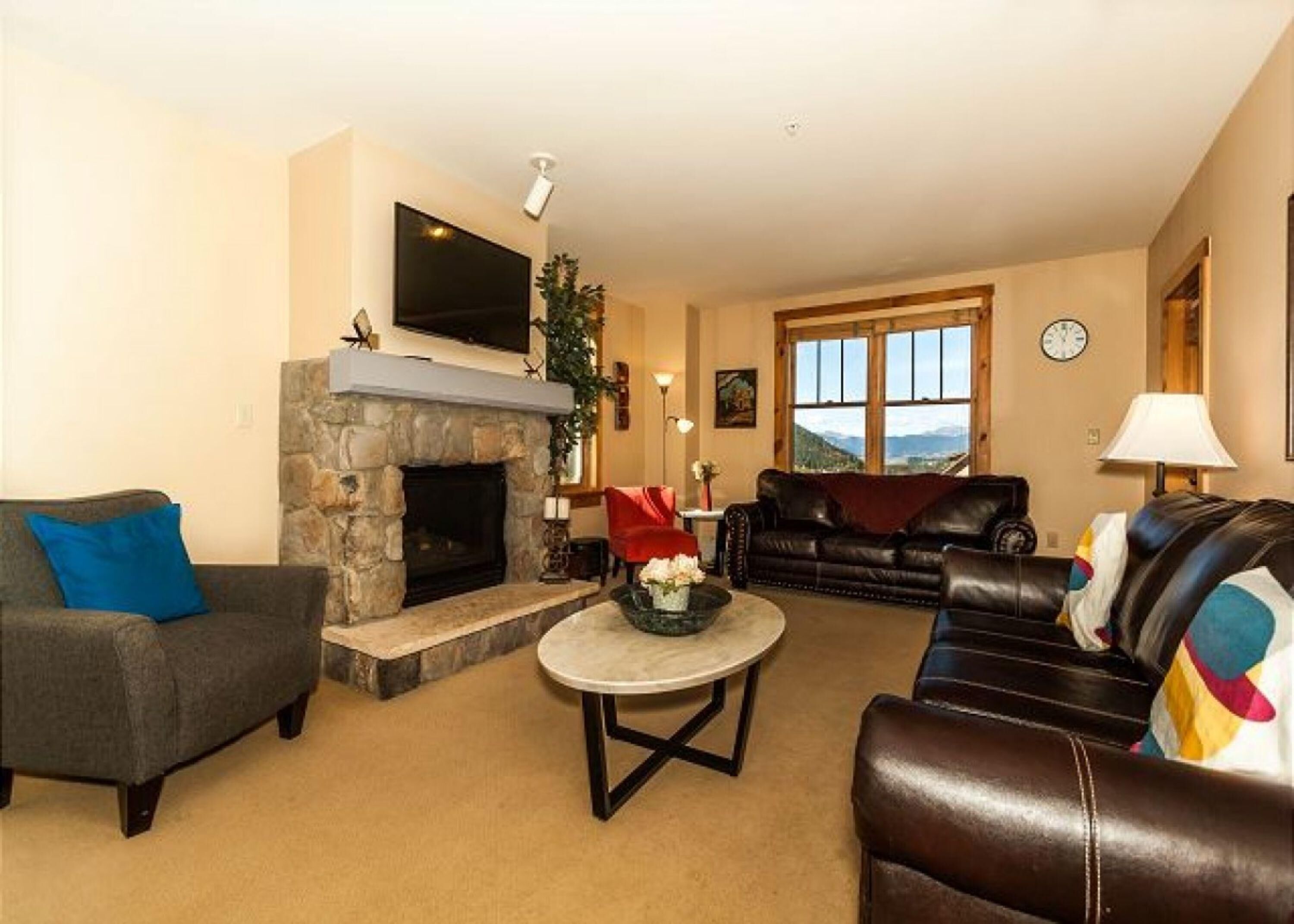 A living room with a stone fireplace, a flat-screen TV mounted above it, a sofa, a coffee table, and a window with a view of the outdoors.