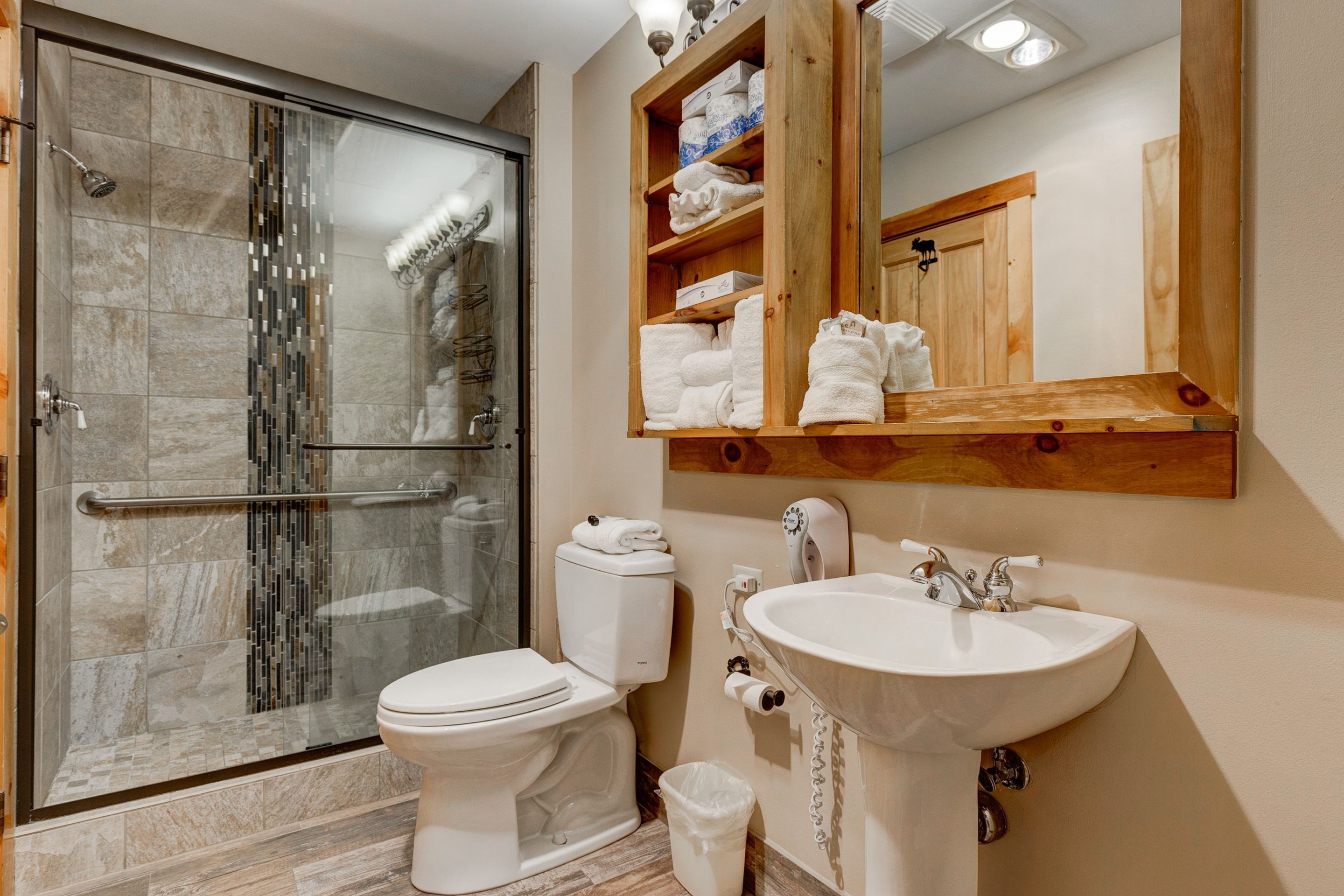 A bathroom with a glass-enclosed shower, a toilet, a sink, and a wooden shelf with towels.