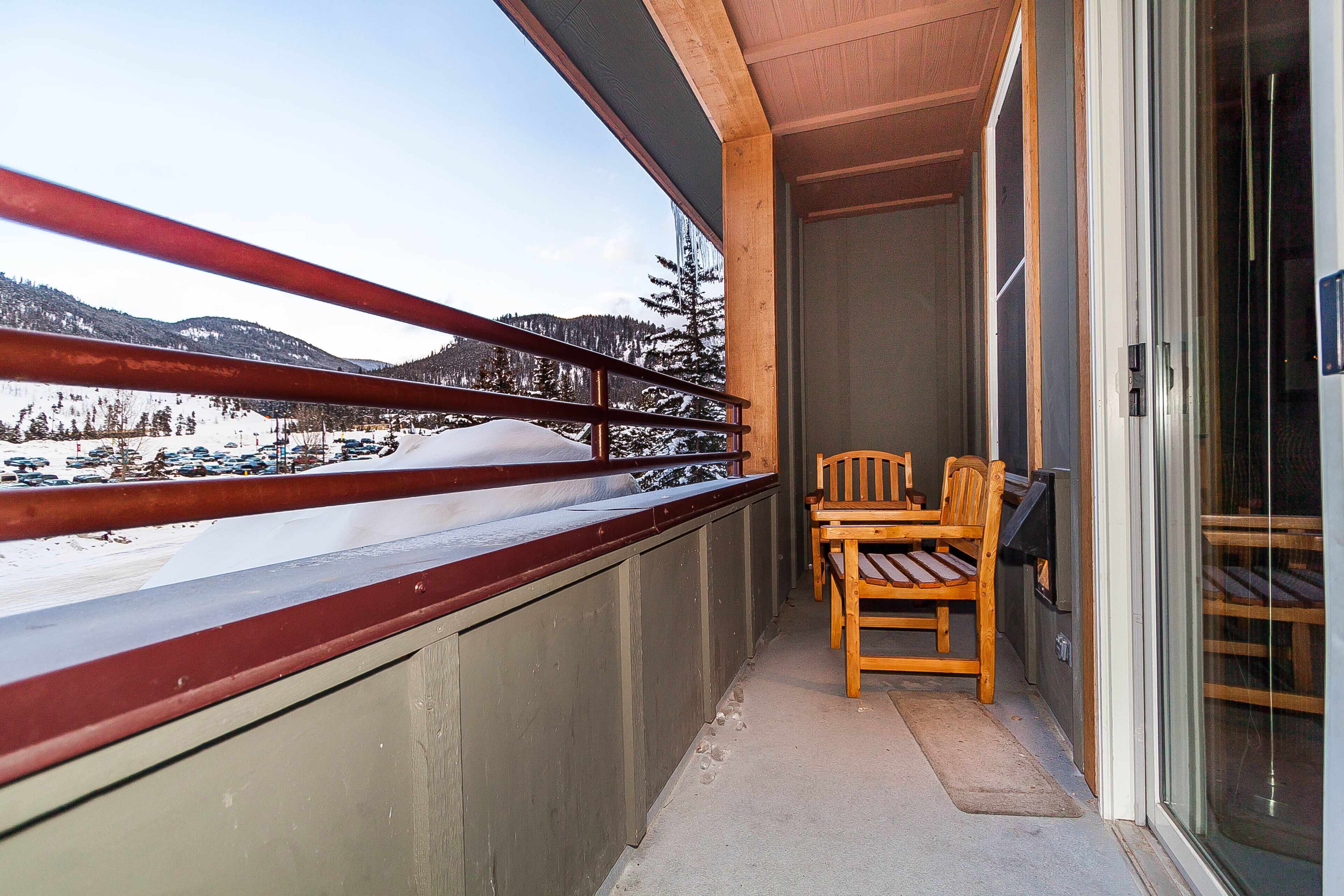 A balcony with a wooden chair and a table, overlooking a snowy landscape with mountains and parked cars.