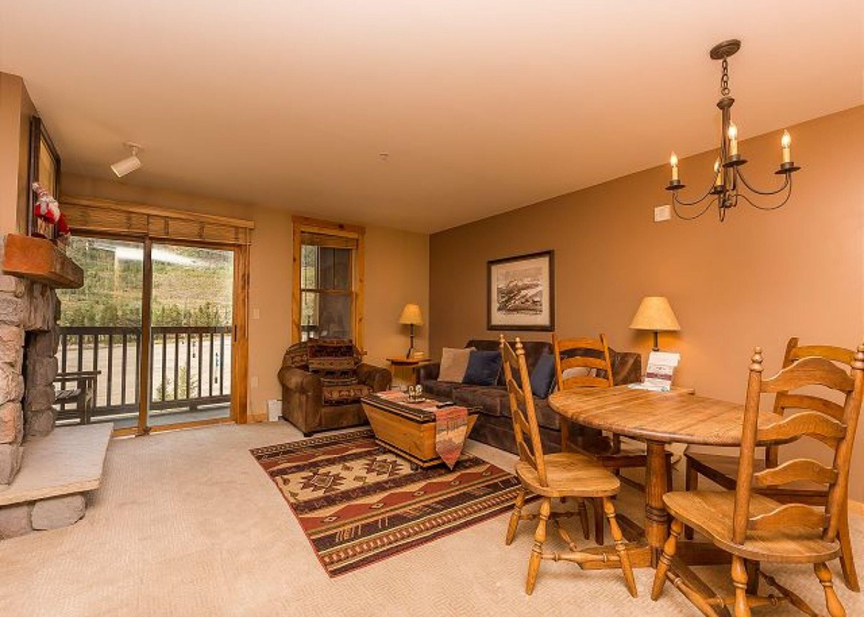 A cozy living room with a fireplace, a dining table with chairs, a sofa, and a large window with a view of the outdoors.