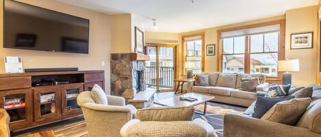 A spacious living room with a flat-screen TV, comfortable seating, a stone fireplace, and large windows offering a view of the outside.