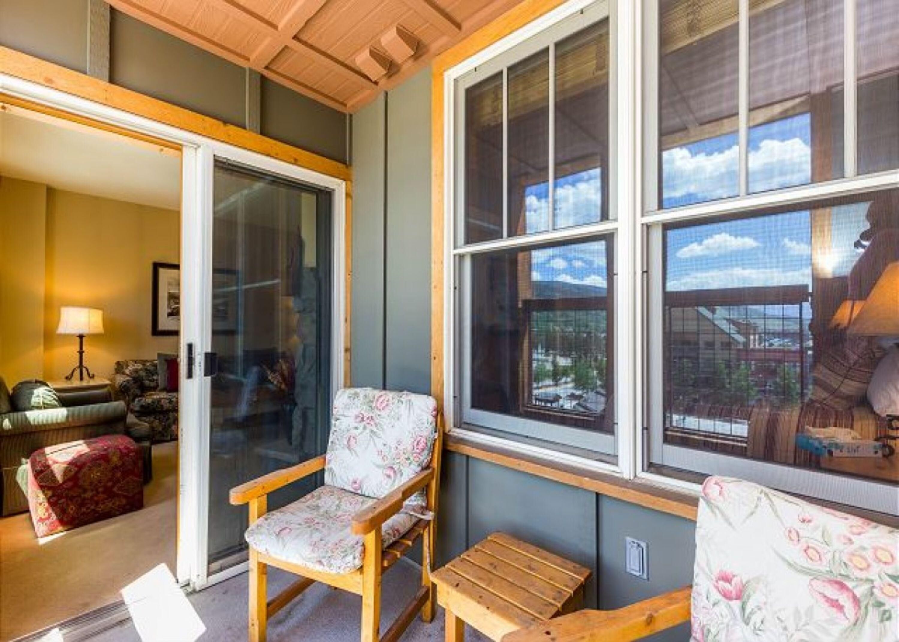 A cozy room with a wooden ceiling, a floral-patterned chair, a wooden table, and large windows offering a view of the outdoors.