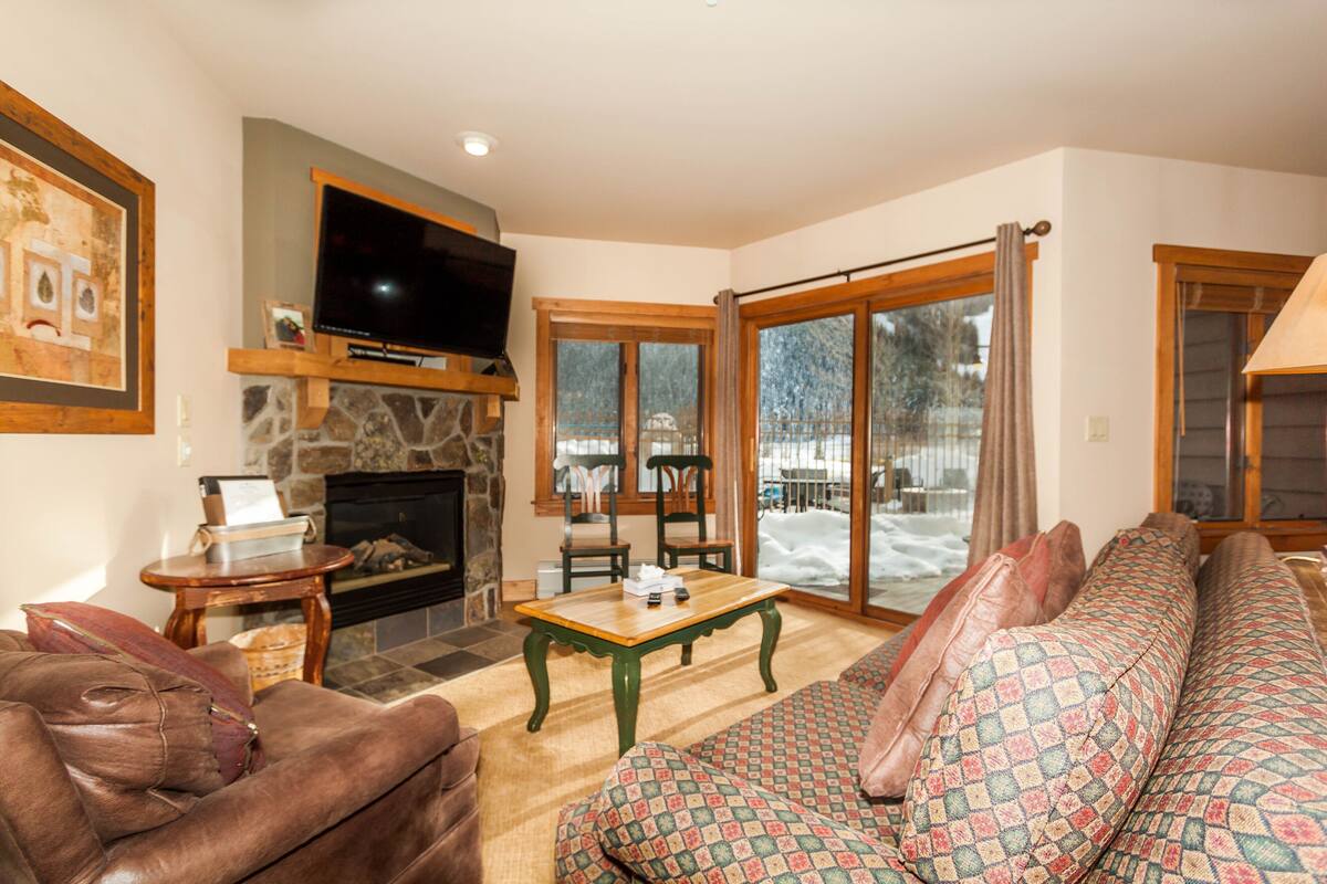A cozy living room with a fireplace, a television, and a large window with a view of a snowy landscape.