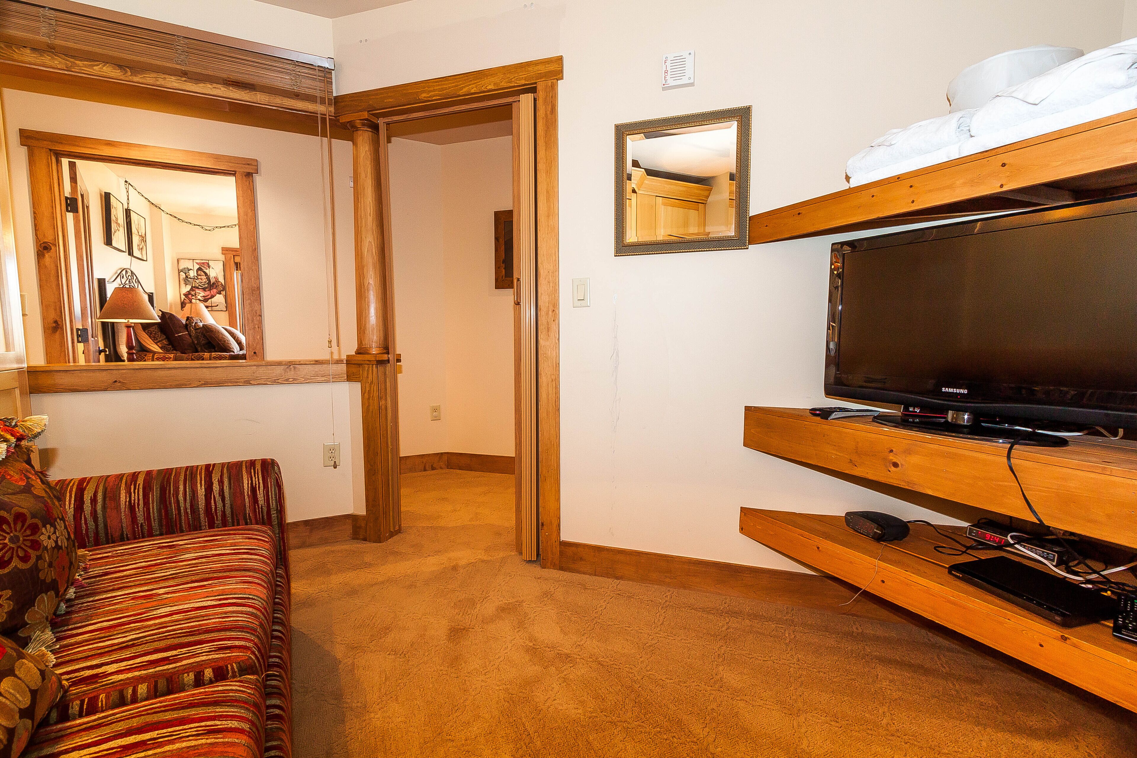 A room with a wooden bench, a sofa, a television on a wooden stand, and a mirror on the wall.