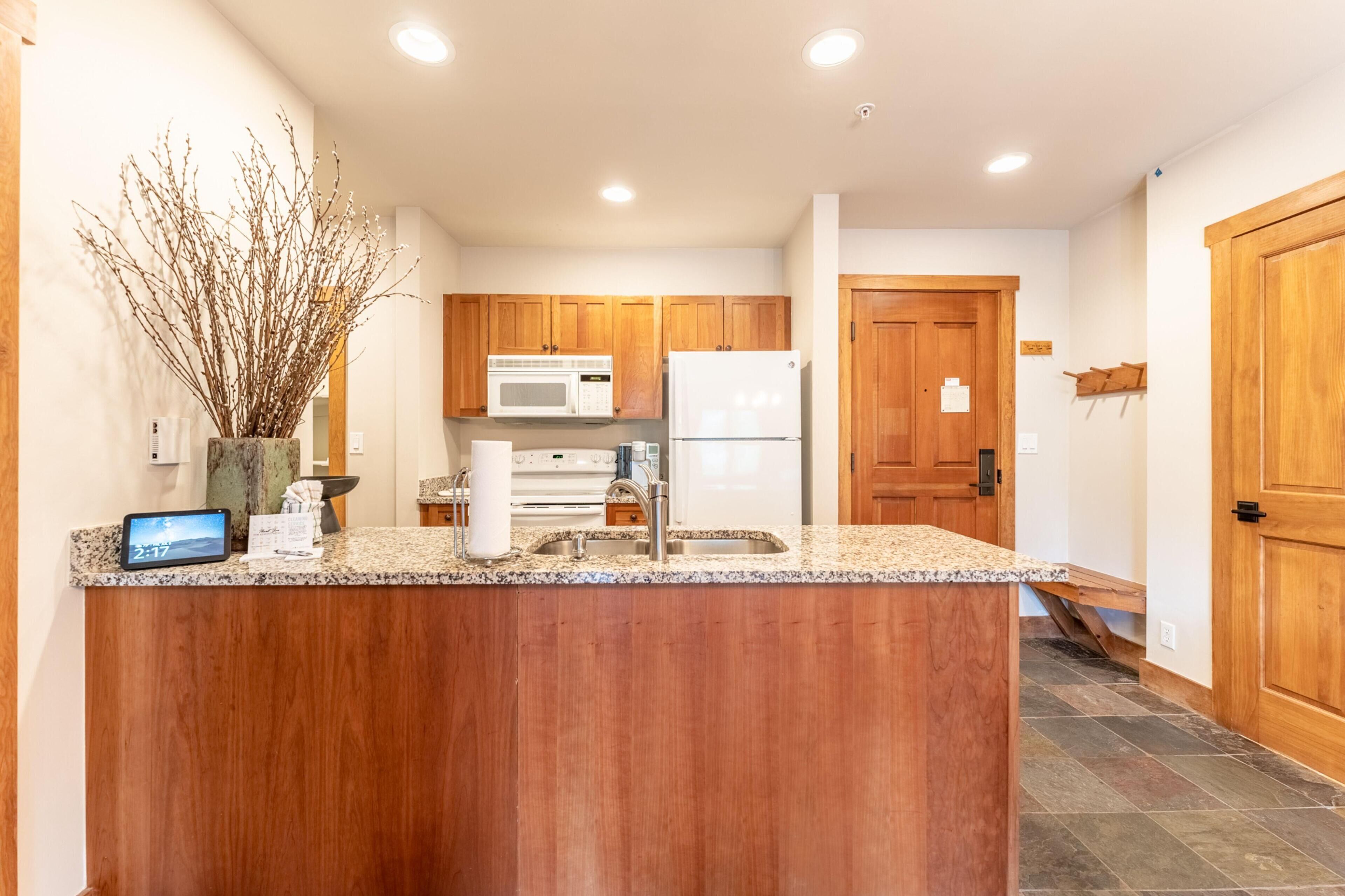 A modern kitchen with wooden cabinets, a granite countertop, a microwave, a refrigerator, and a sink.