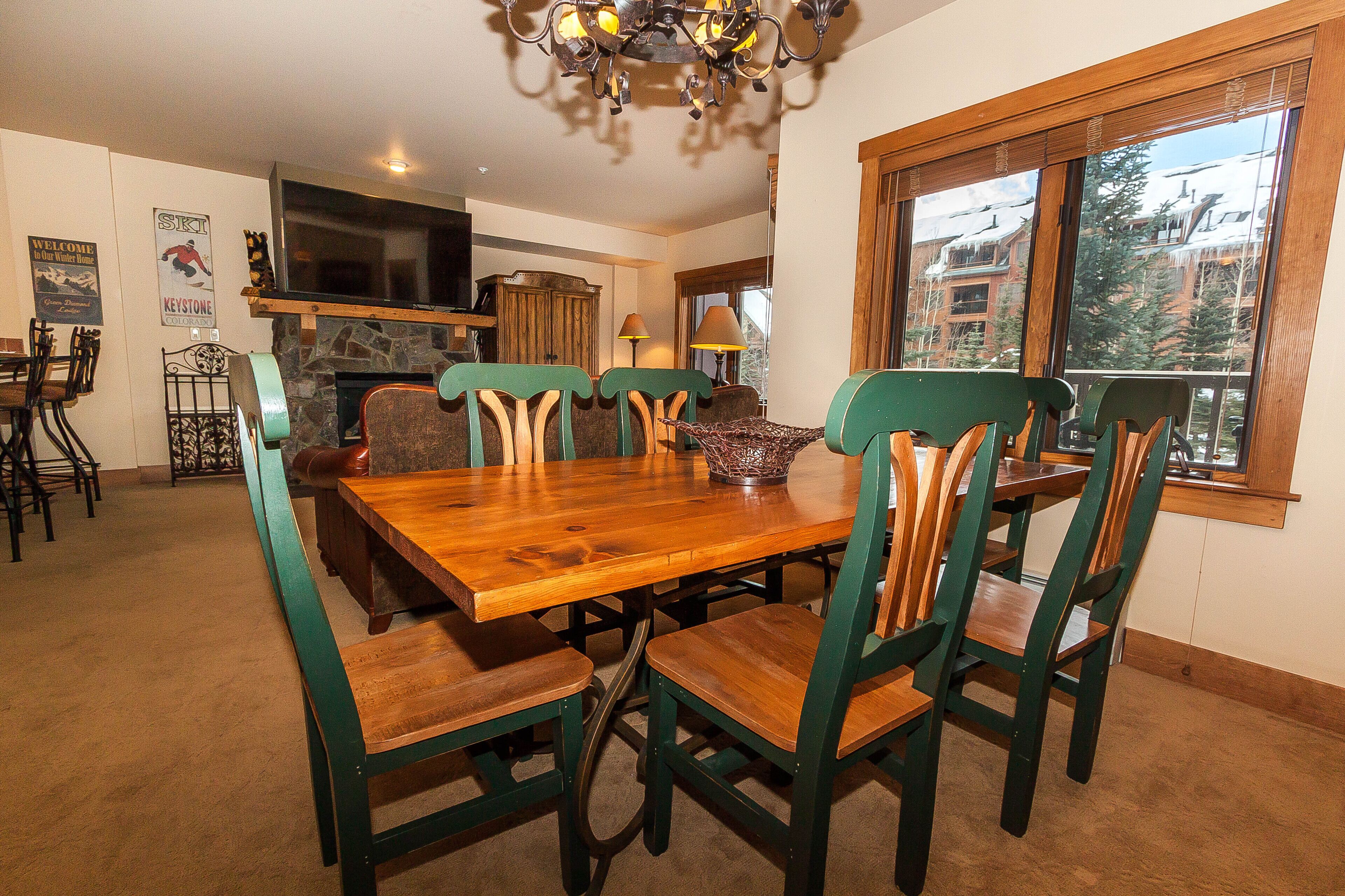 A dining area with a wooden table and green chairs, a stone fireplace, and a large window with a view of buildings.