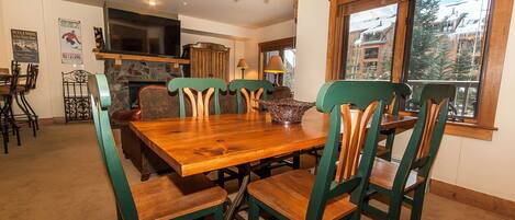 A dining area with a wooden table and green chairs, a stone fireplace, and a large window with a view of buildings.
