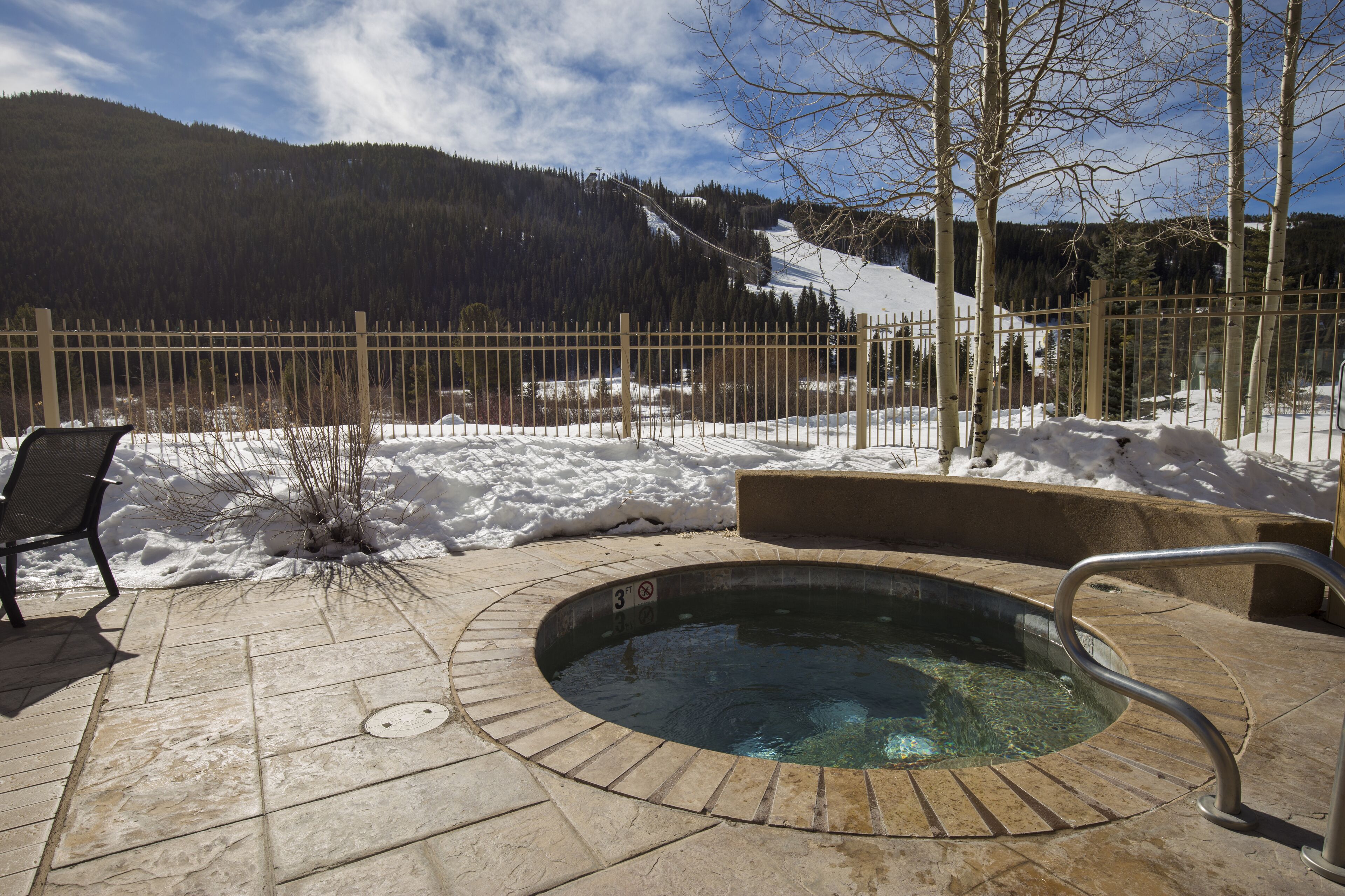 A hot tub with a stone surround, a wooden fence, and a chair in a snowy outdoor area with a mountain view.