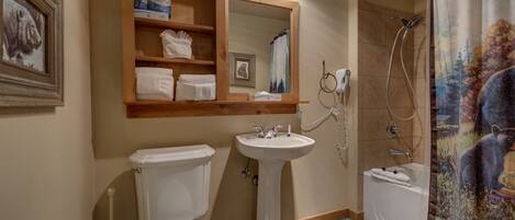 A bathroom with a toilet, sink, and shower. There is a mirror above the sink with shelves holding towels and toiletries.