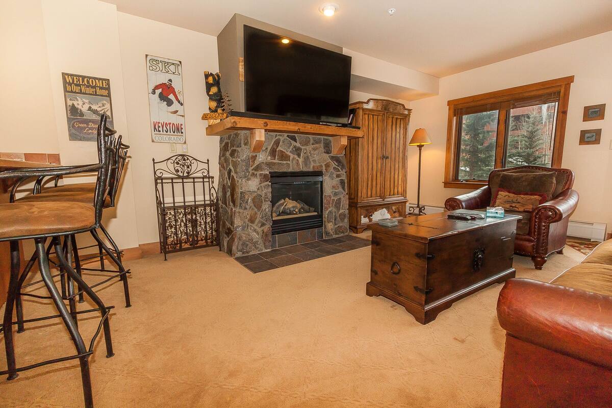 A living room with a stone fireplace, a television mounted above it, and a cozy seating area with a leather armchair and a wooden coffee table.