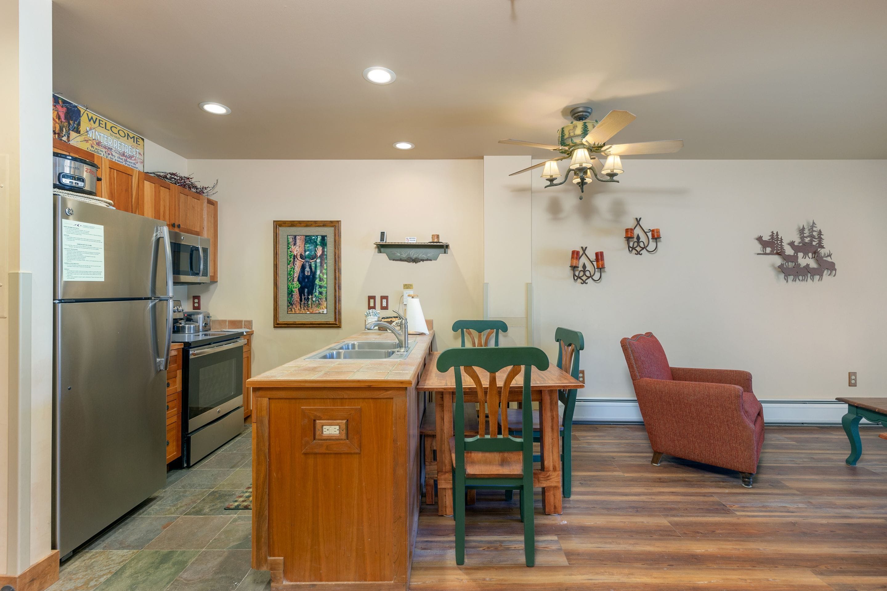 A kitchen with a refrigerator, microwave, oven, and sink. There is a dining area with a wooden table and chairs. A ceiling fan and wall decorations are visible.