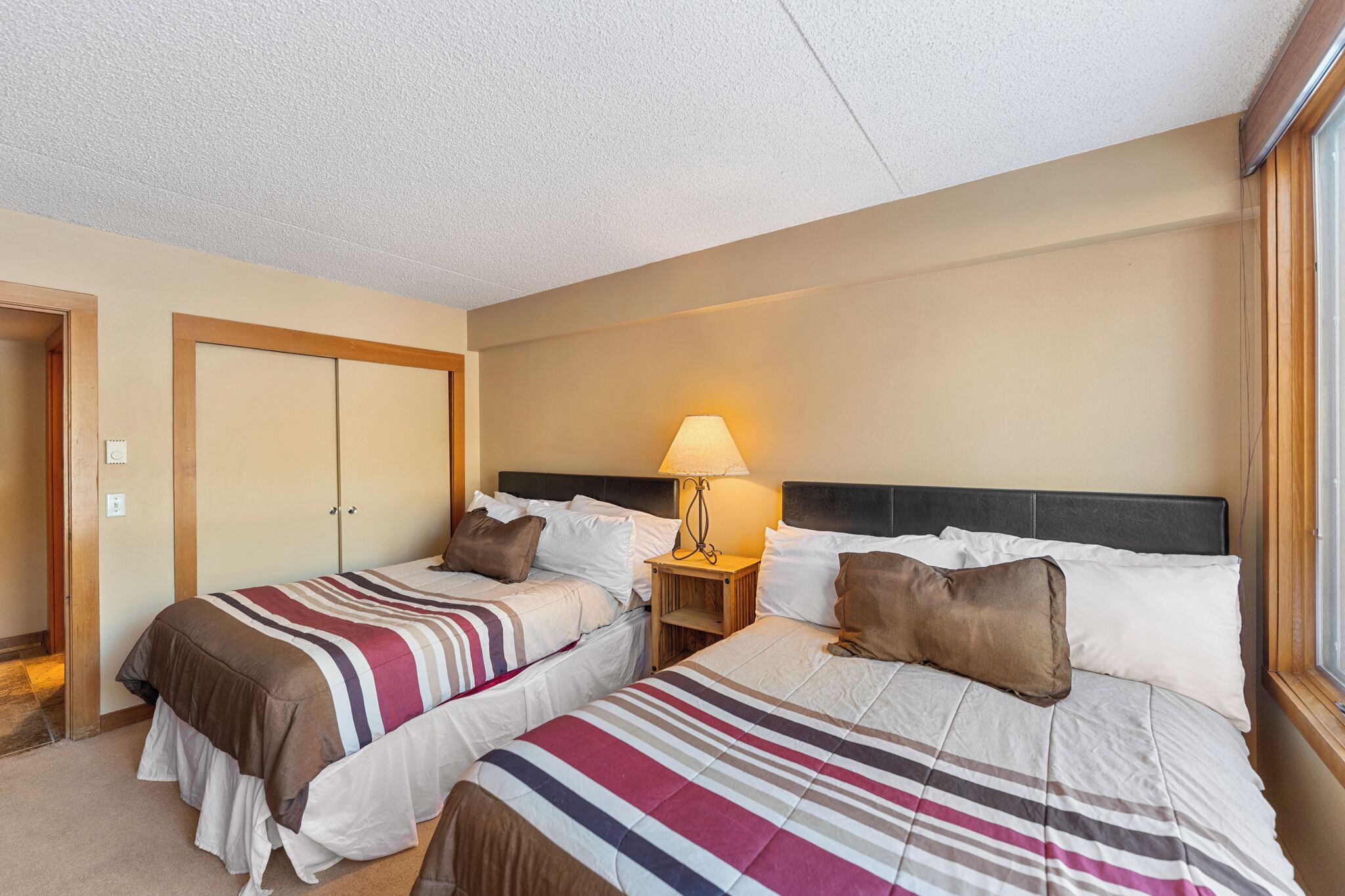 A hotel room with two beds, a nightstand with a lamp, and a door.