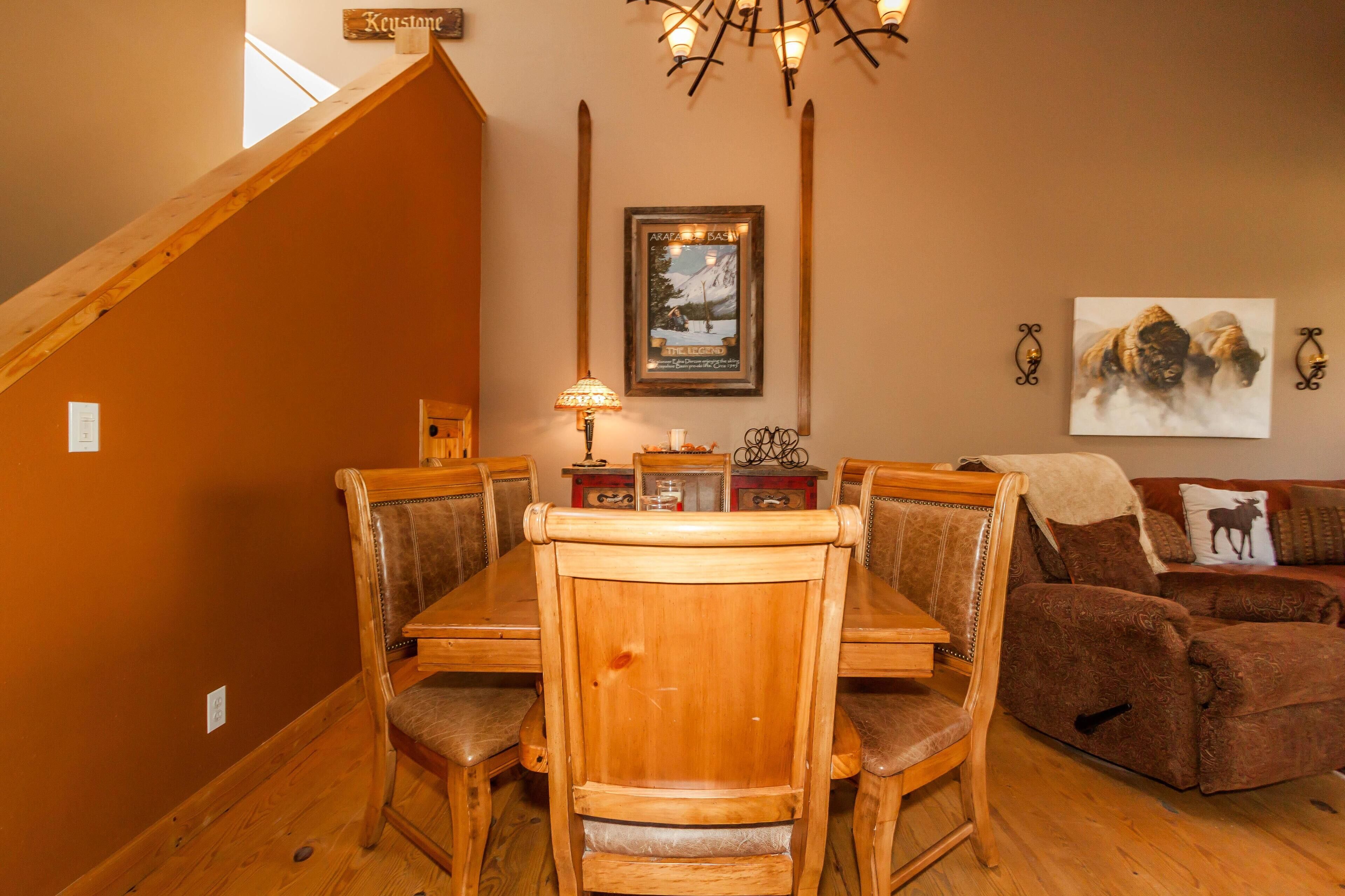 A dining area with a wooden table and chairs, a chandelier, a framed picture on the wall, and a couch with a cushion.