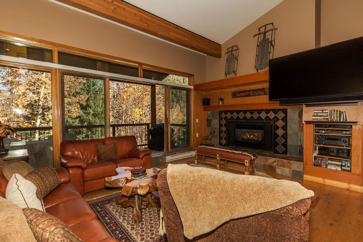 A cozy living room with a fireplace, leather sofa, and large windows overlooking a forest.
