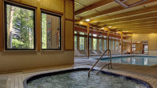 Une grande piscine intérieure avec de larges baies vitrées, des poutres en bois et un sol carrelé.
