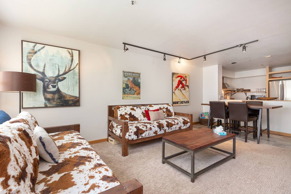 A living room with a cowhide-patterned sofa, a wooden coffee table, and a dining area with a kitchen in the background.
