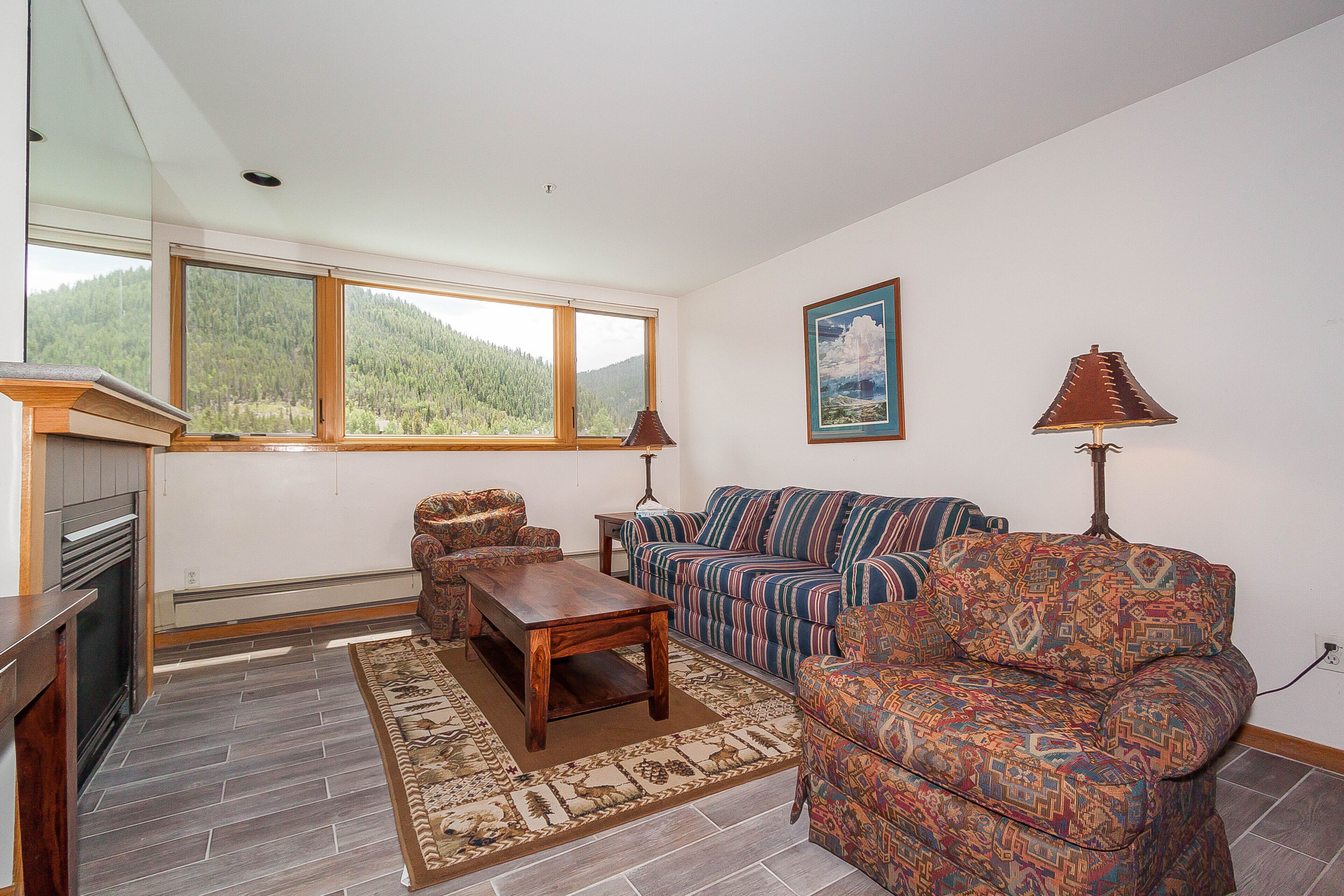 A living room with a fireplace, a sofa, a patterned rug, and a view of a forest through large windows.