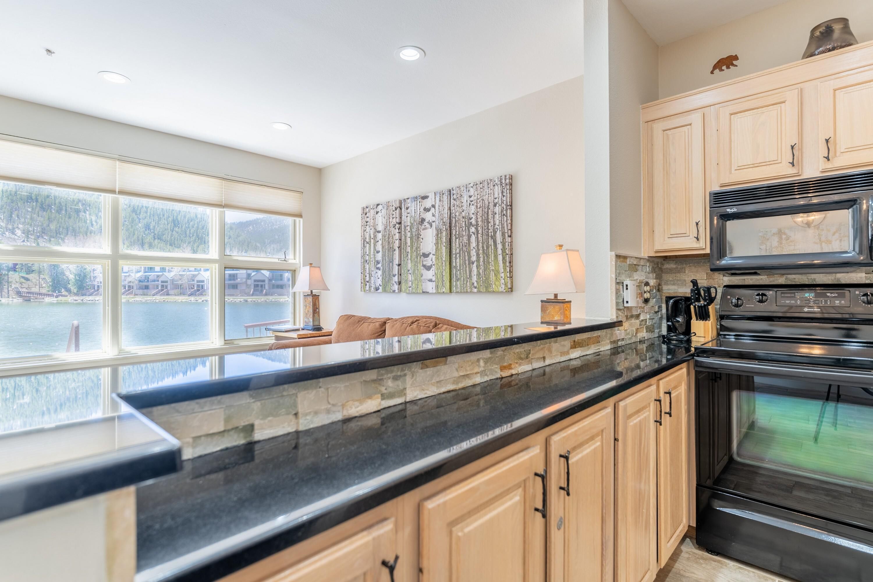 A kitchen with a large window, stone countertop, wooden cabinets, and a stainless steel oven.