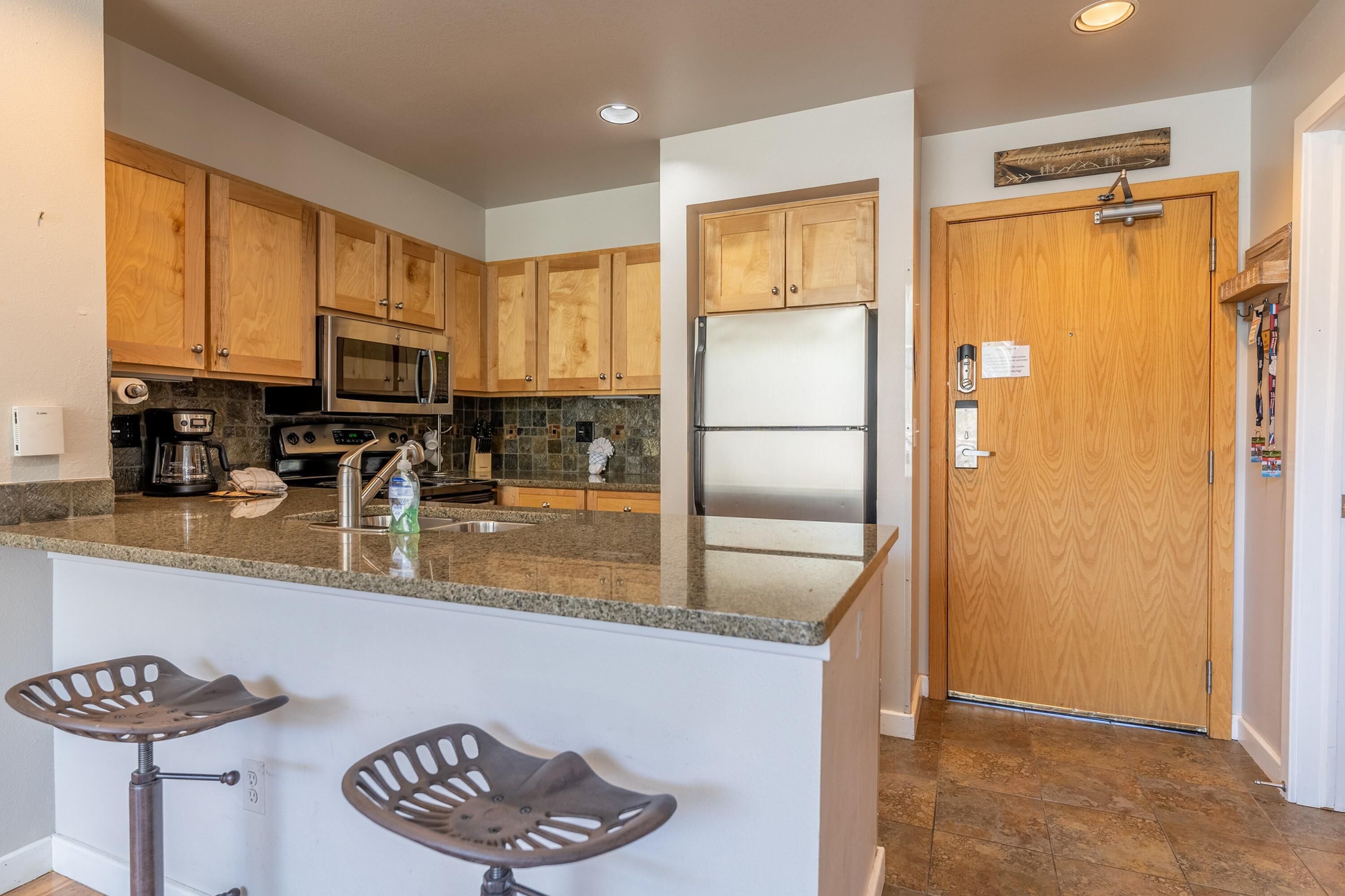 A kitchen with wooden cabinets, stainless steel appliances, a granite countertop, and two bar stools.