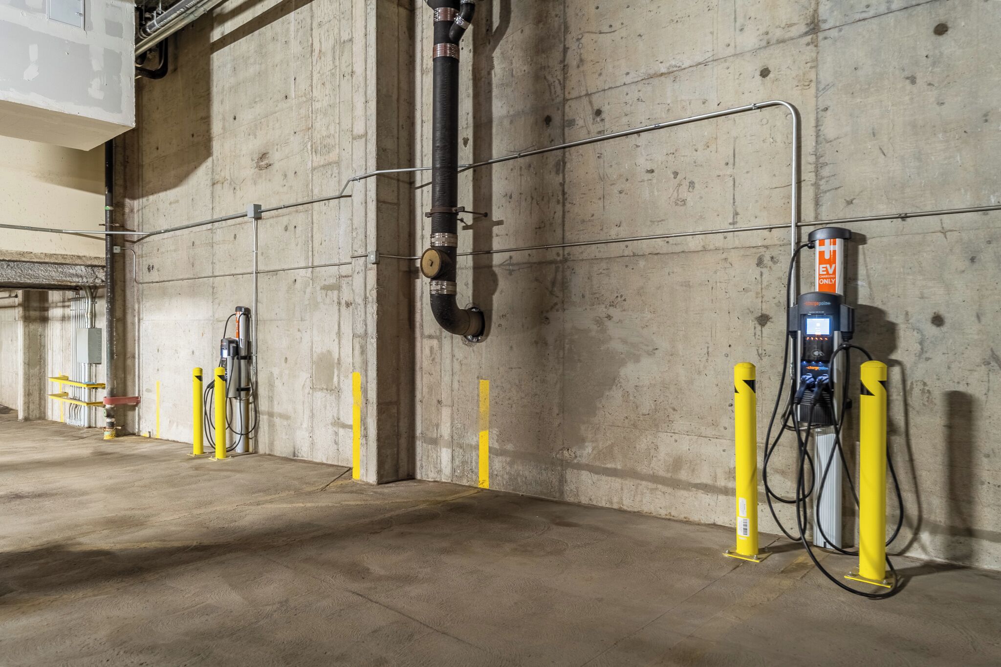 Un espace de stationnement intérieur avec des murs en béton, des bornes jaunes et des bornes de recharge pour véhicules électriques.