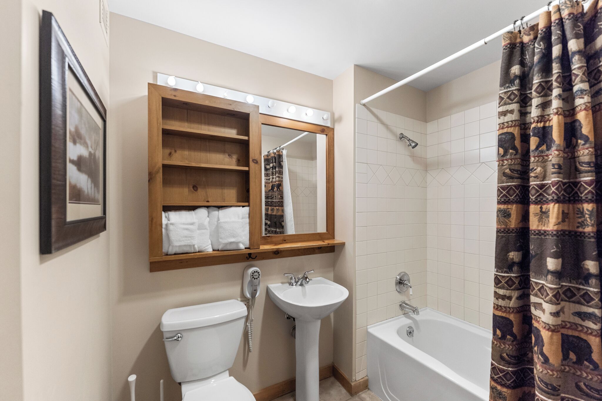 Ein Badezimmer mit Toilette, Waschbecken und Badewanne, einem Holzboden über dem Waschbecken, einem beleuchteten Spiegel und einer Duschvorhang mit Muster.