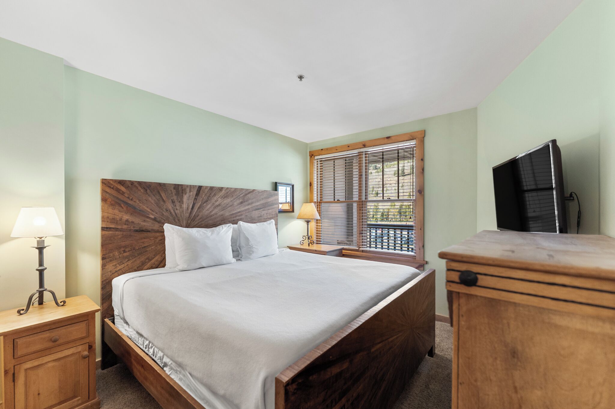 A bedroom with a wooden headboard, a bed with white linens, a nightstand with a lamp, a wooden dresser, a window with blinds, and a television on the wall.