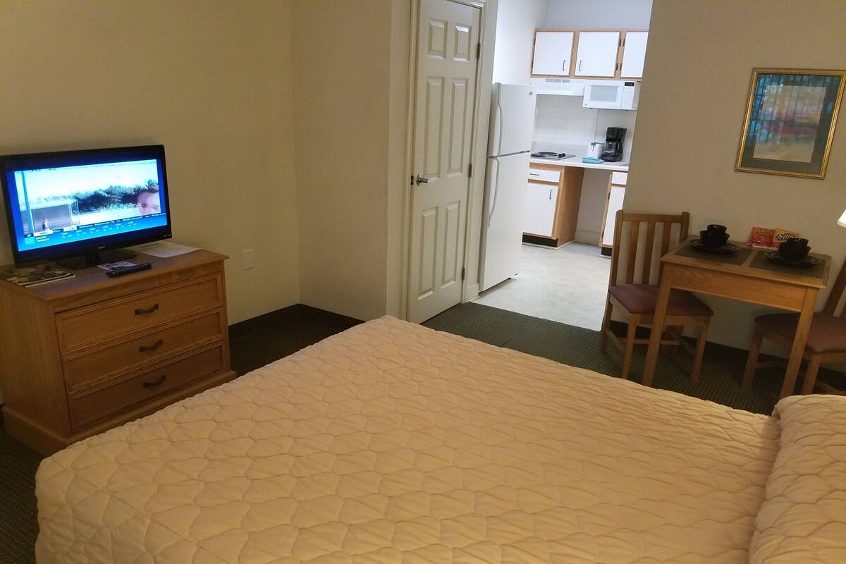 A hotel room with a bed, a TV on a wooden dresser, a desk with a chair, and a kitchenette in the background.