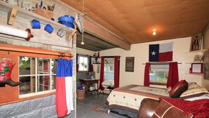 Una habitación con una cama, techo de madera, una bandera de Texas y una ventana con cortinas rojas.