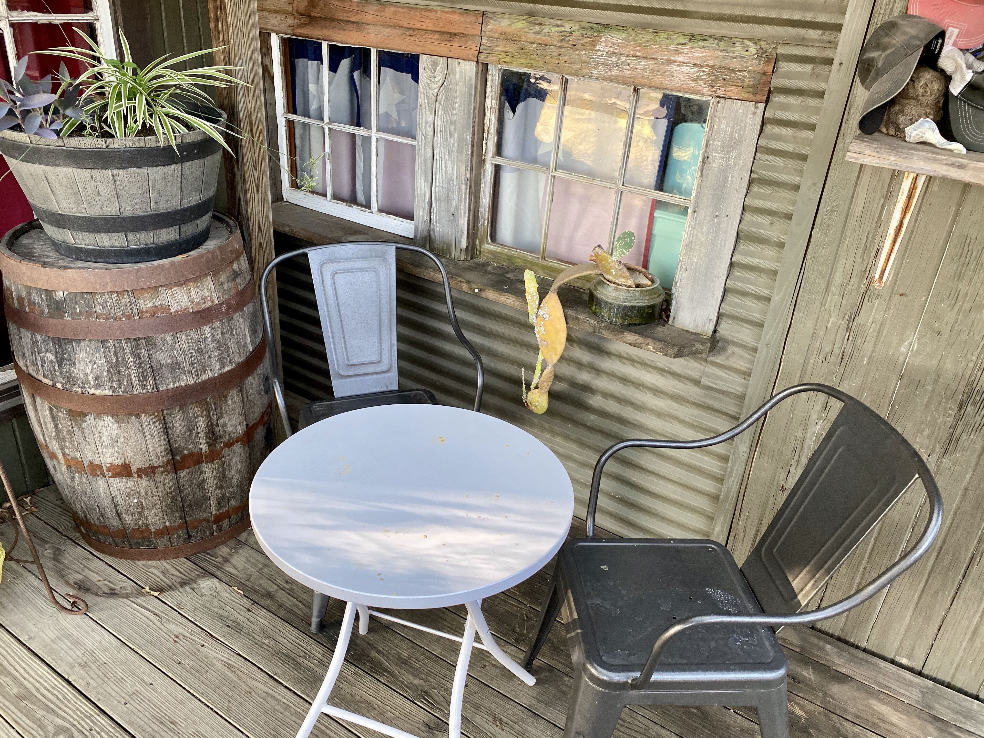 A rustic outdoor seating area with a metal table and chairs, a large wooden barrel, and a window with curtains.
