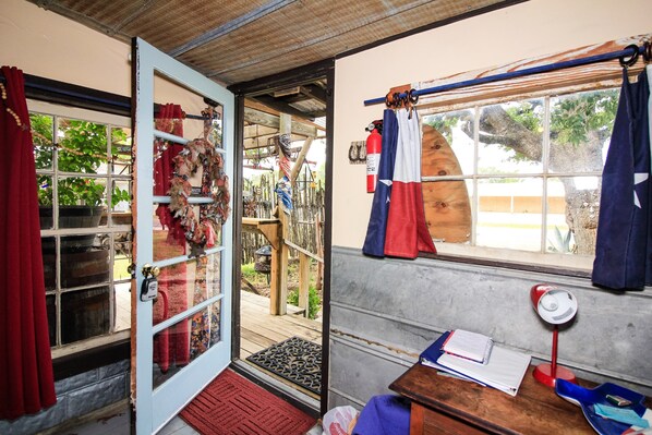 A room with a wooden door, a window with curtains, a desk with a lamp, and a view of the outside.