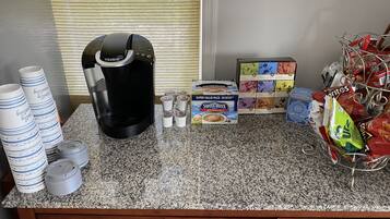 A kitchen counter with a coffee maker, stacks of paper cups, a box of Swiss Miss tea bags, and a basket of snacks.
