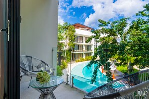 Uma varanda com mesa de vidro, cadeiras de vime e vista para uma piscina e um edifício com varandas.