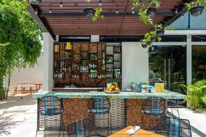 Área de bar ao ar livre com balcão, cadeiras e uma variedade de garrafas e frutas.