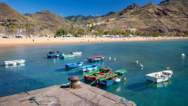 Escena costera con barcos amarrados en un muelle, una playa de arena y montañas al fondo.