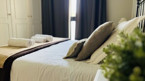 Cama bien hecha con sábanas blancas y dos almohadas, una toalla enrollada al lado, y una ventana con cortinas oscuras.